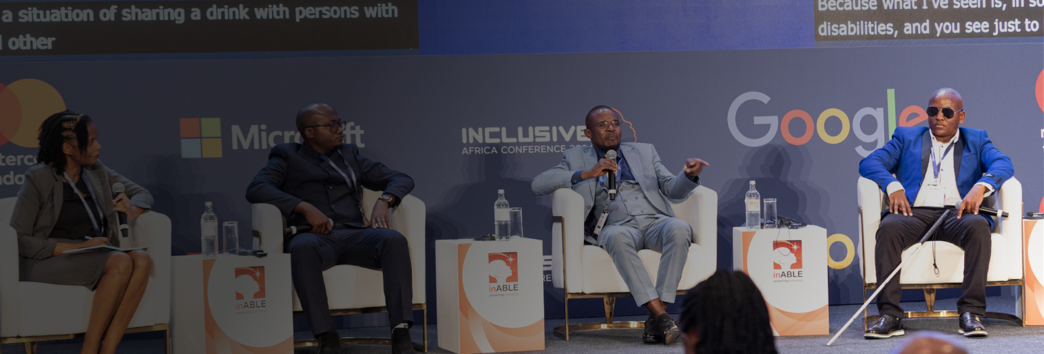 Mask group (11) A panel discussion at the Inclusive Africa Conference 2024. Three male panelists and one female moderator are seated on stage in white chairs, engaging in conversation. The background features sponsor logos including Mastercard Foundation, Microsoft, Google, and inABLE. The panelists are dressed in formal attire; one man on the far right uses a white cane, indicating visual impairment. An audience is visible in the foreground, attentively listening to the discussion. The conference branding and date are prominently displayed on the backdrop.