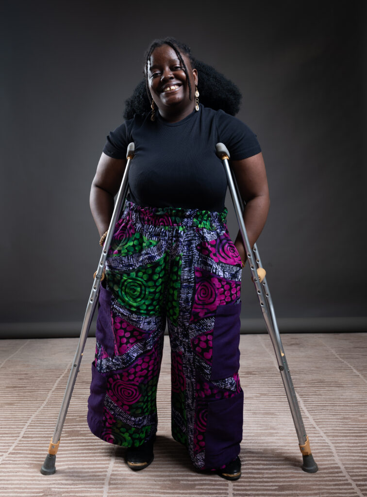 Pelemo stands confidently using crutches, dressed in a black short-sleeve top and colorful, vibrant patchwork pants featuring green, pink, purple, and white patterns. She has long, curly hair and wears dangling earrings. The background is a simple, dark studio setting with a beige patterned floor.