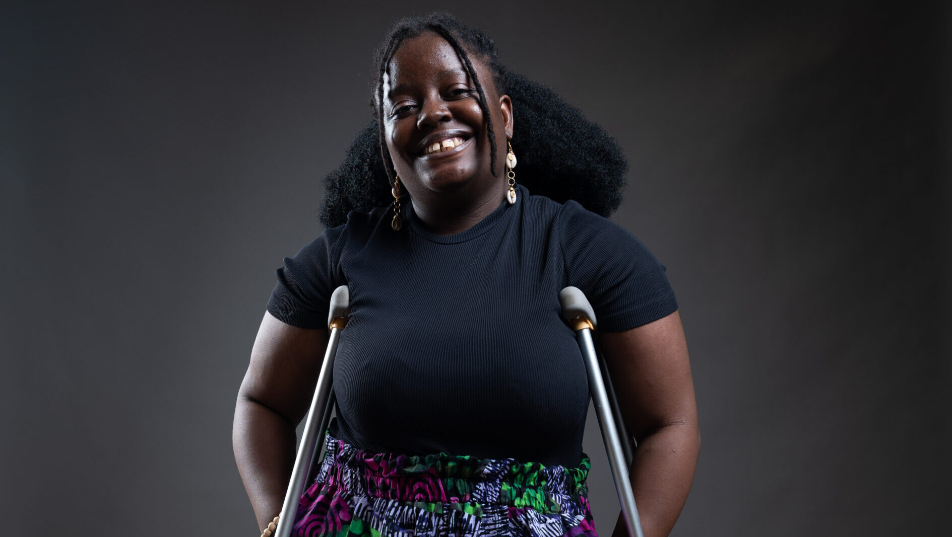 Pelemo stands confidently using crutches, dressed in a black short-sleeve top and colorful, vibrant patchwork pants featuring green, pink, purple, and white patterns. She has long, curly hair and wears dangling earrings. The background is a simple, dark studio setting with a beige patterned floor.