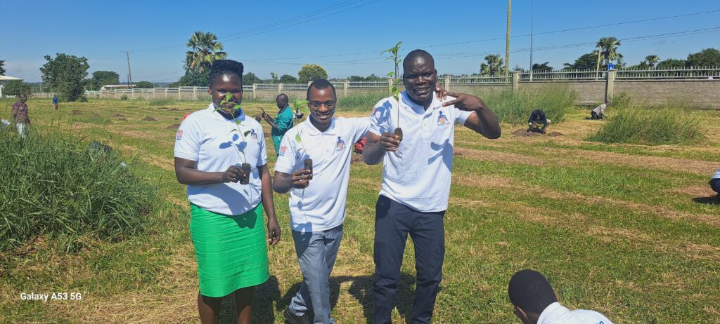 Three people including Harunah standing outdoors on a sunny day, holding young tree seedlings in small soil bags, with others planting in the background. They are wearing white polo shirts with logos and appear to be participating in a tree-planting activity on a grassy field.