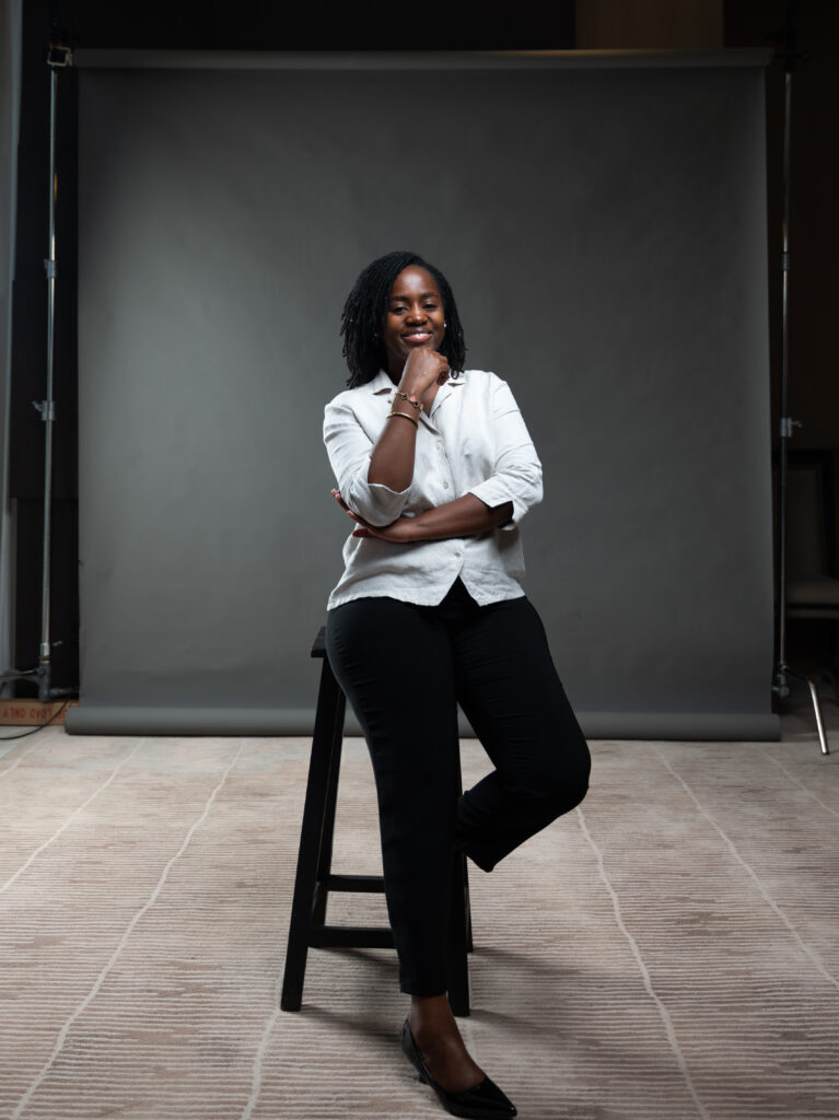 A photo of Doreck, with dreadlocks sitting on a stool, smiling and looking directly at the camera. She is wearing a light-colored, button-up shirt and black pants, and has her chin resting on one hand. Her other arm is crossed over her chest. The background is a plain, dark gray backdrop with a hint of a studio floor and equipment visible at the edges.