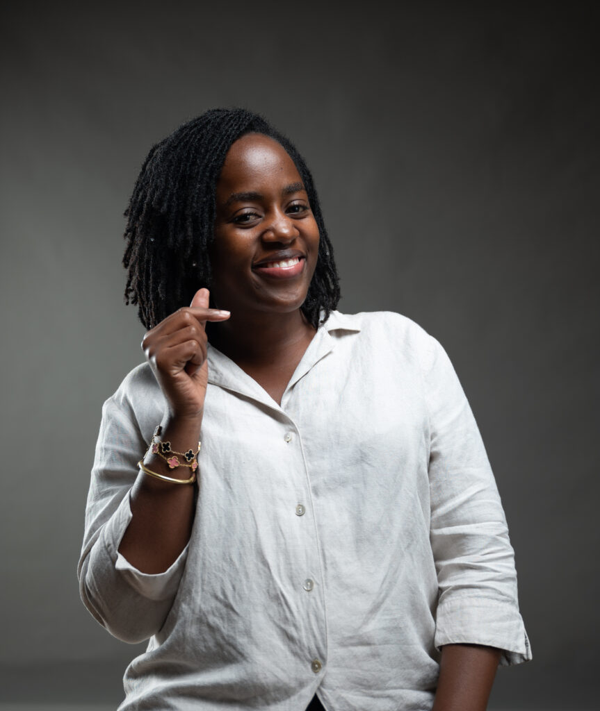 A photo of Doreck with dreadlocks is shown from the chest up, smiling at the camera. She is wearing a light-colored, button-up shirt and a bracelet with a pink floral design. She is holding her thumb and index finger together to form a small heart shape. The background is a plain, dark gray color.