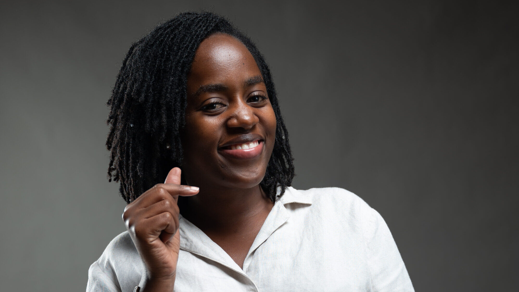 A photo of Doreck with dreadlocks is shown from the chest up, smiling at the camera. She is wearing a light-colored, button-up shirt and a bracelet with a pink floral design. She is holding her thumb and index finger together to form a small heart shape. The background is a plain, dark gray color.