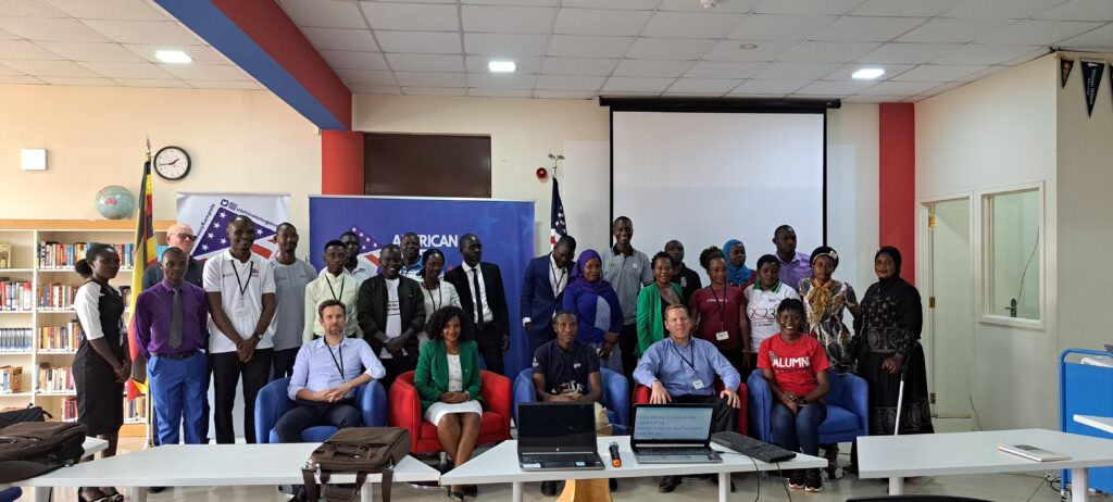 Group photo of diverse participants, including Harunah, men and women, gathered indoors at an event in a library setting with banners reading 'American' in the background. Some are seated in front, while others stand behind, with flags, bookshelves, and a projection screen visible in the room."
