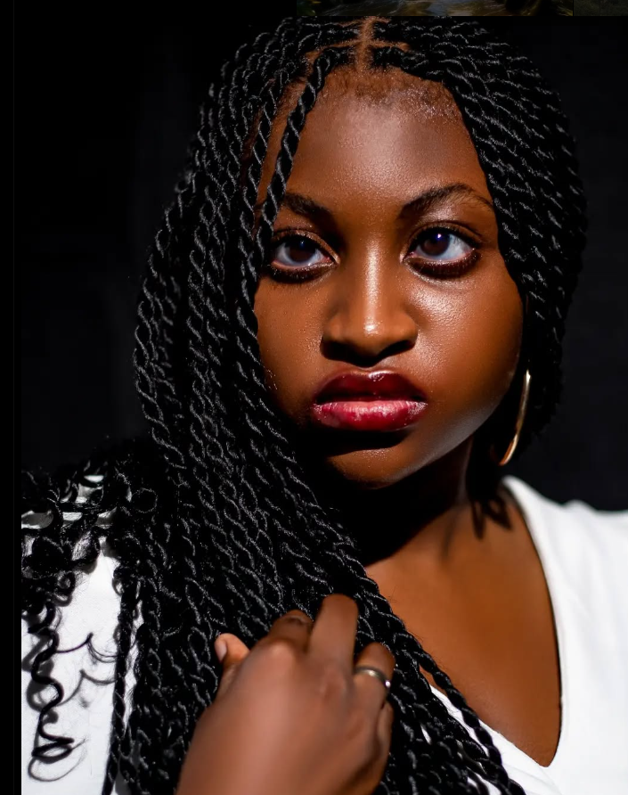 A close-up portrait of Alvarita looking to her right, her eyes wide. She has twist braids that frame her face and fall over her shoulder. She is wearing a white shirt and a gold hoop earring. She has on a ring on her right hand. The background is a dark, shadowy black.