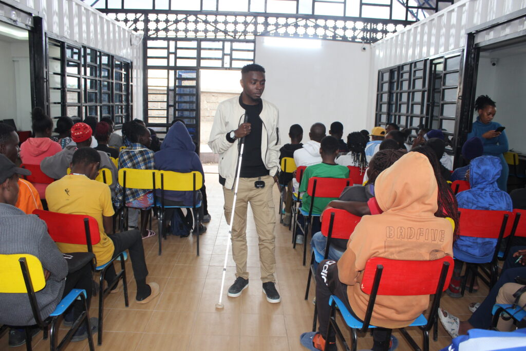 Julius is holding his white cane and standing inside Isinya Digital Hub. He speaks to a seated group in a brightly lit classroom. Attendees, wearing casual attire, focus on the speaker with attentive expressions.