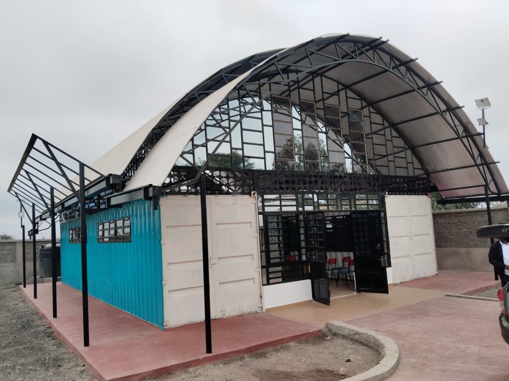 A picture of Isinya Digital Hub, a modern building with a curved metal roof, glass windows, and white and blue walls. The design is industrial and innovative against a cloudy sky.