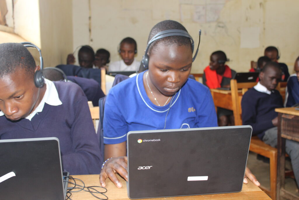 
A low-angle shot shows a group of students sitting at wooden desks in a classroom, each using a laptop and wearing headphones with a microphone. In the foreground, a girl in a blue t-shirt with "80s" on the sleeve looks intently at her laptop. Her hands are on the keyboard, and she has a headset on. To her left, a boy in a navy blue sweater with a white collared shirt looks down at his laptop. Other students are visible in the background. The classroom walls are a dirty white, and the desks are old and scratched.