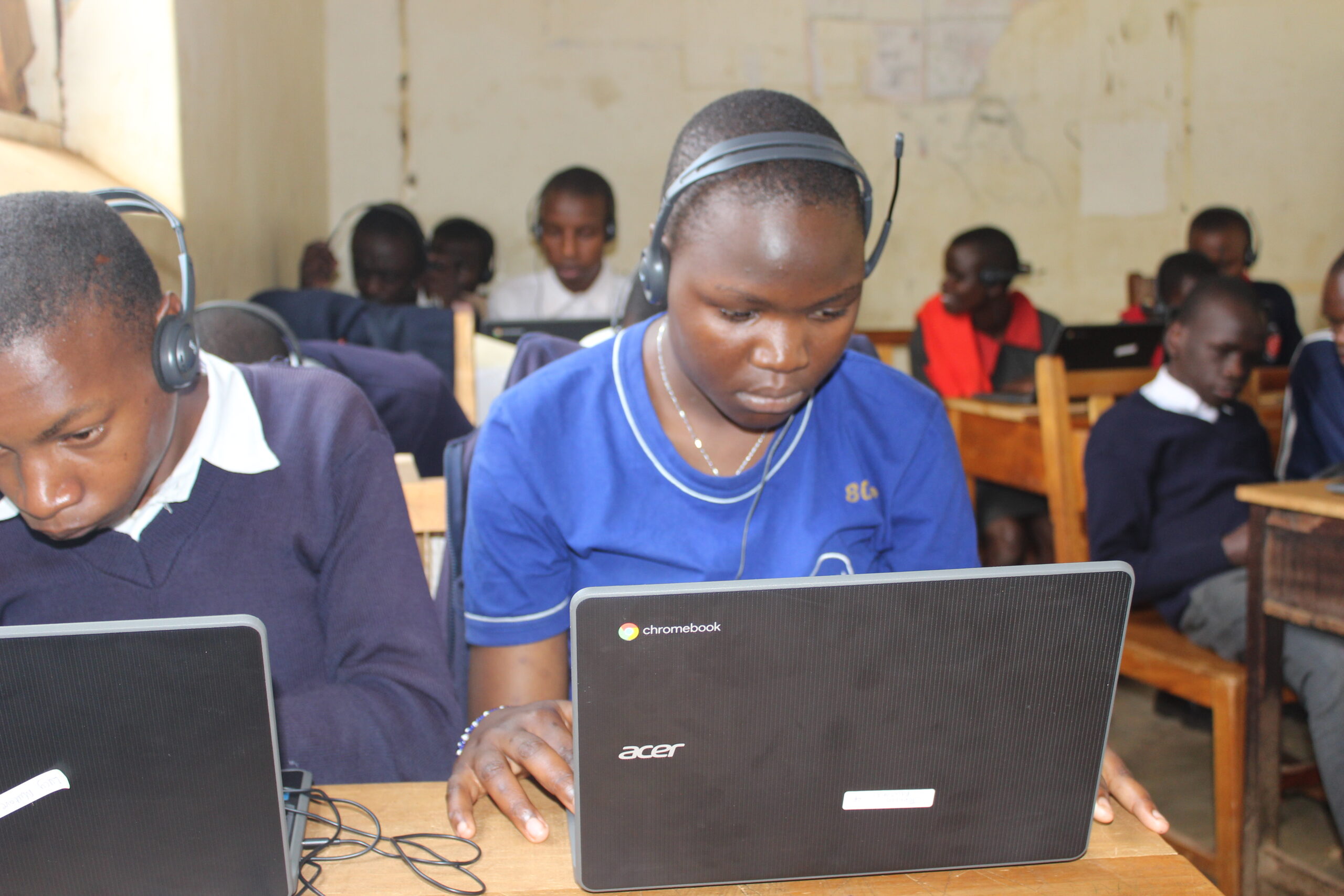 A low-angle shot shows a group of students sitting at wooden desks in a classroom, each using a laptop and wearing headphones with a microphone. In the foreground, a girl in a blue t-shirt with "80s" on the sleeve looks intently at her laptop. Her hands are on the keyboard, and she has a headset on. To her left, a boy in a navy blue sweater with a white collared shirt looks down at his laptop. Other students are visible in the background. The classroom walls are a dirty white, and the desks are old and scratched.