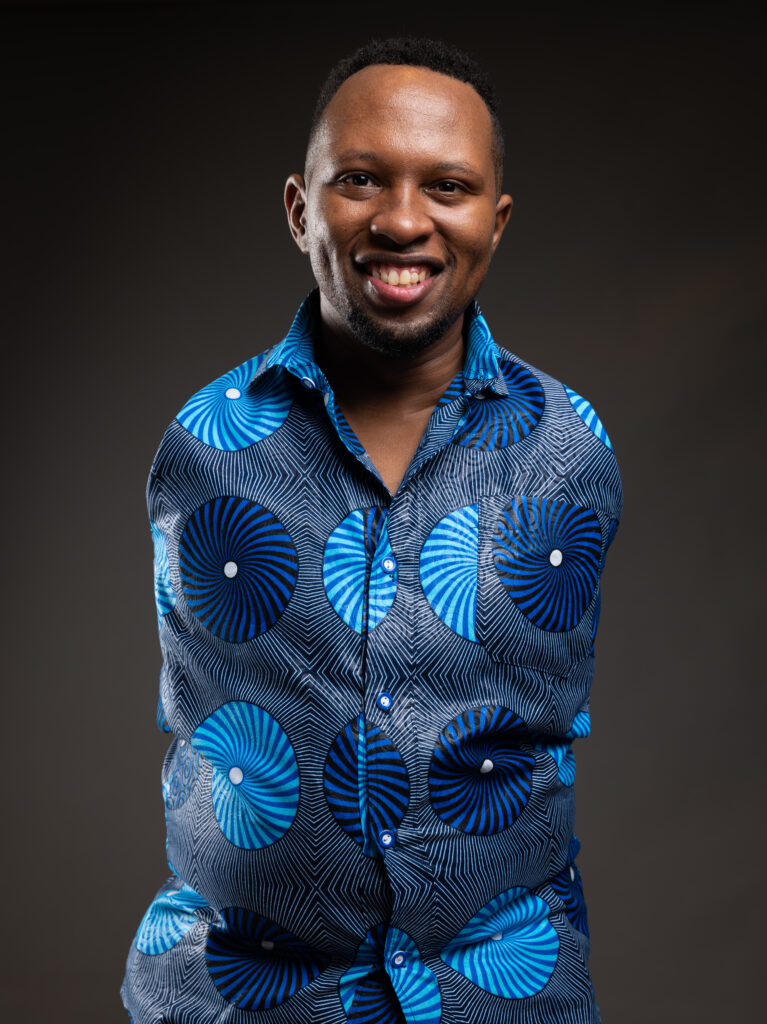A picture of Sammy wearing a blue patterned shirt with geometric circles stands against a dark background.
