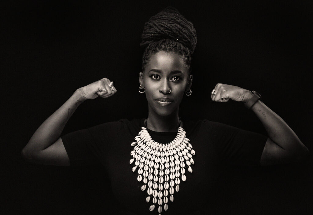 A low-angle, black and white studio portrait of Nadia flexing her biceps and looking directly at the camera with a subtle smirk. Her hair is in dreadlocks, pulled up into a large bun on top of her head. She wears small hoop earrings, a black t-shirt, and a large statement necklace made of dozens of white, shell-like pieces. The background is a solid, dark gray.