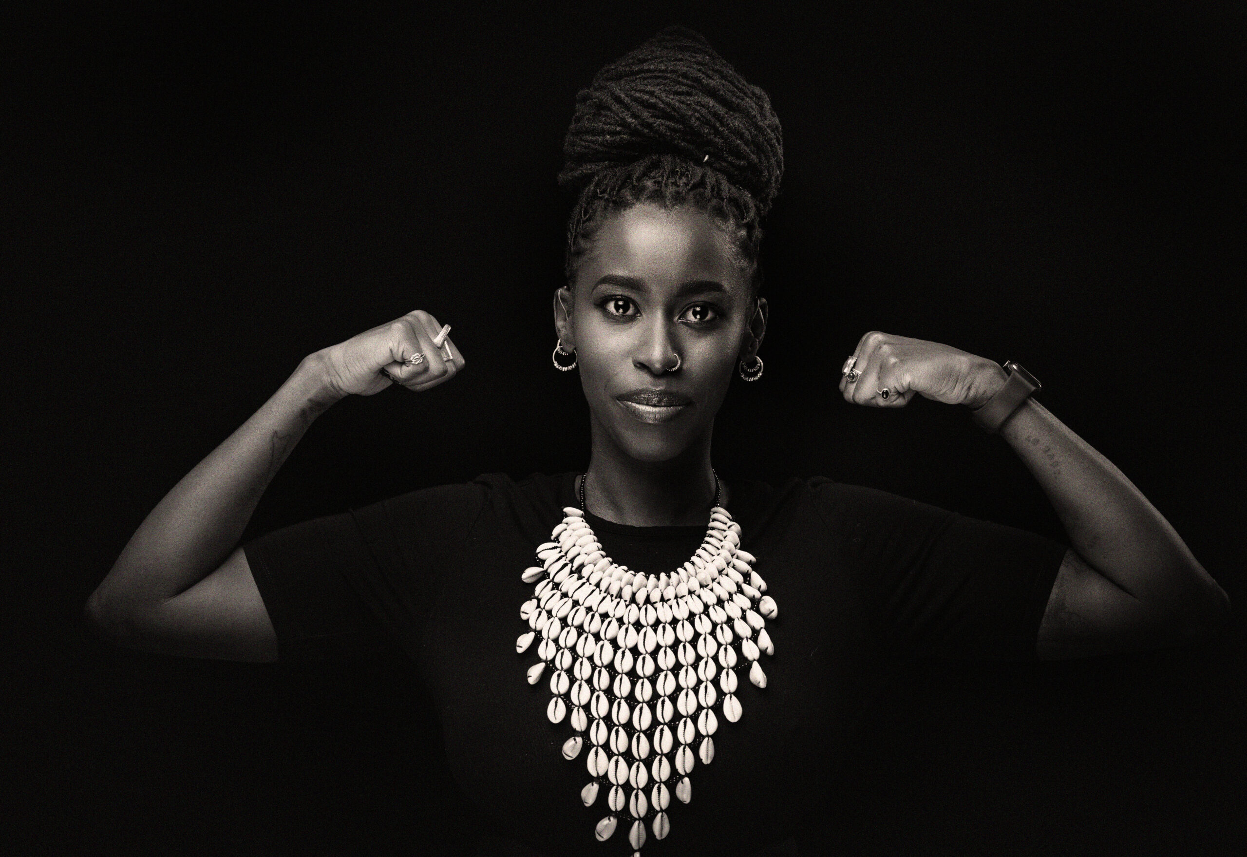 A low-angle, black and white studio portrait of Nadia flexing her biceps and looking directly at the camera with a subtle smirk. Her hair is in dreadlocks, pulled up into a large bun on top of her head. She wears small hoop earrings, a black t-shirt, and a large statement necklace made of dozens of white, shell-like pieces. The background is a solid, dark gray.