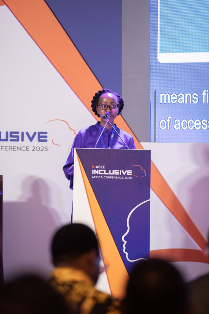 A medium shot of Angela speaking at a podium. She is wearing a purple collared shirt and glasses. Her hair is in cornrows and she holds the microphone with both hands, looking down slightly. The podium has the orange and white logo for the "Inclusive Africa Conference 2025." Behind her is a large screen with text, and a large banner with the same logo. The foreground is blurry, showing the back of an audience member's head.