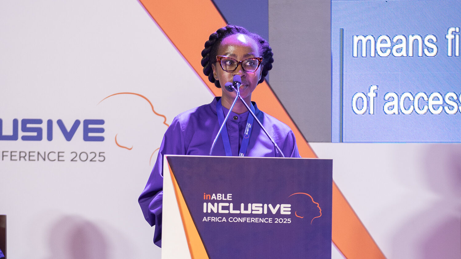 A medium shot of Angela speaking at a podium. She is wearing a purple collared shirt and glasses. Her hair is in cornrows and she holds the microphone with both hands, looking down slightly. The podium has the orange and white logo for the "Inclusive Africa Conference 2025." Behind her is a large screen with text, and a large banner with the same logo. The foreground is blurry, showing the back of an audience member's head.