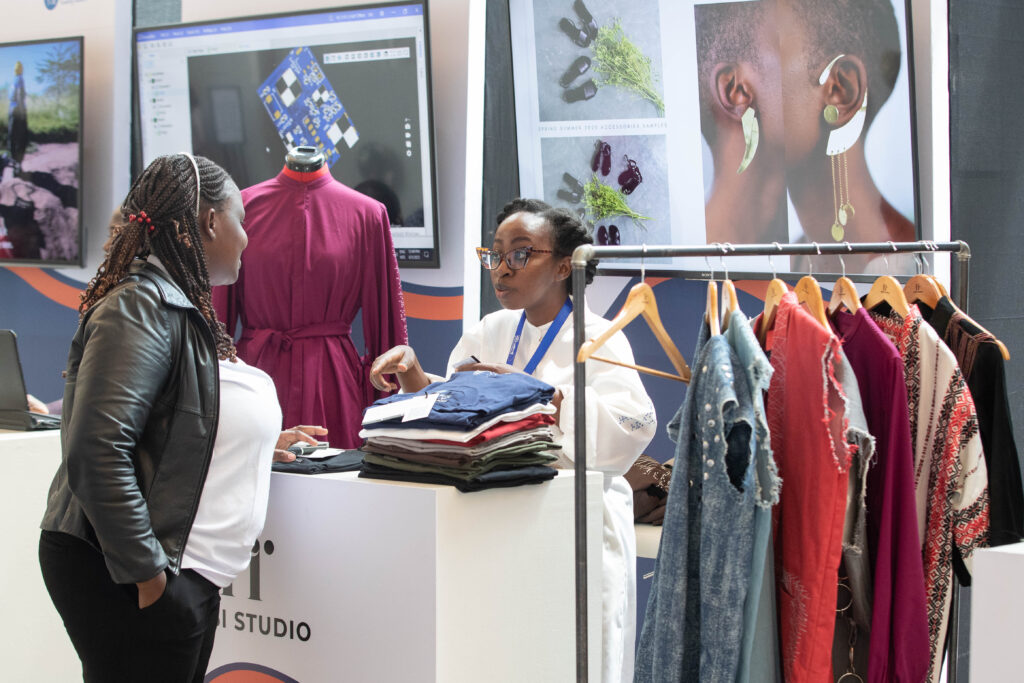 Angela is wearing glasses and an employee ID badge. She is speaking with a female attendee across a white booth with the text "HISI STUDIO" on the front. The employee, wearing a white shirt, is gesturing towards a stack of folded blue shirts on the counter. The female attendee is wearing a black leather jacket over a white top, is leaning on the counter and listening. To the right of the booth, a rack of clothes is displayed, and behind the two women, a mannequin wears a magenta dress. In the background, a large screen displays a design program, and a poster shows a close-up of a person with a stylish earring.