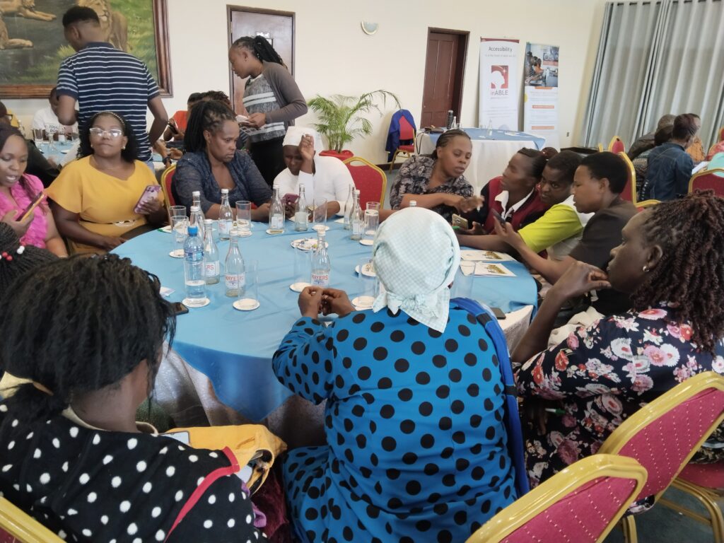 A group of people seated around round tables covered with blue tablecloths, engaged in conversation during a workshop or event. Bottles of water and glasses are placed on the tables. Some participants are looking at their phones, while others are actively discussing. In the background, banners and posters related to accessibility and inclusion by inABLE and Kilimanjaro Blind Trust Africa are visible.