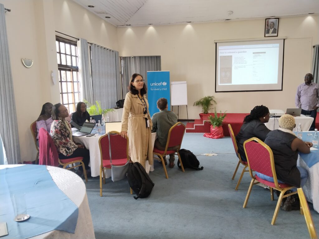 A bright room with a light blue carpet and white walls. A woman in a long beige coat stands in the middle, holding a microphone, with a UNICEF logo visible behind her. Several people are seated at round tables with light blue and white tablecloths. A white screen with text is projected on the back wall, and a man in a plaid shirt stands next to it.