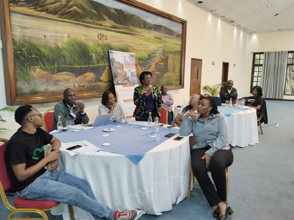 Participants seated around tables during a workshop session. Tabie Kioko (Safaricom) standing and holding a microphone is speaking to the group, while others listen and engage. Laptops, documents, and water bottles are placed on the tables. Behind her, a banner on employability skills and a large mural of lions in the wild are visible on the wall.