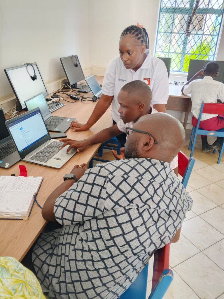 A female instructor wearing a white polo shirt with a logo is leaning over a wooden desk to help a young male student and an adult man seated at a laptop computer. The student is also looking at the screen and pointing, and the adult man, wearing a black and white patterned shirt, is sitting with his back to the camera, wearing glasses. Several other laptops and monitors are visible on the desk and in the background, suggesting a computer lab or classroom setting. Two other students are partially visible at desks further back in the room.