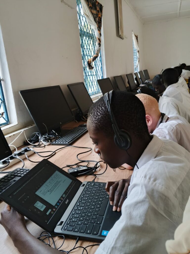 A student wearing headphones works closely on a laptop in a computer lab, with several other students beside him and unused desktop monitors lined up on the desk.