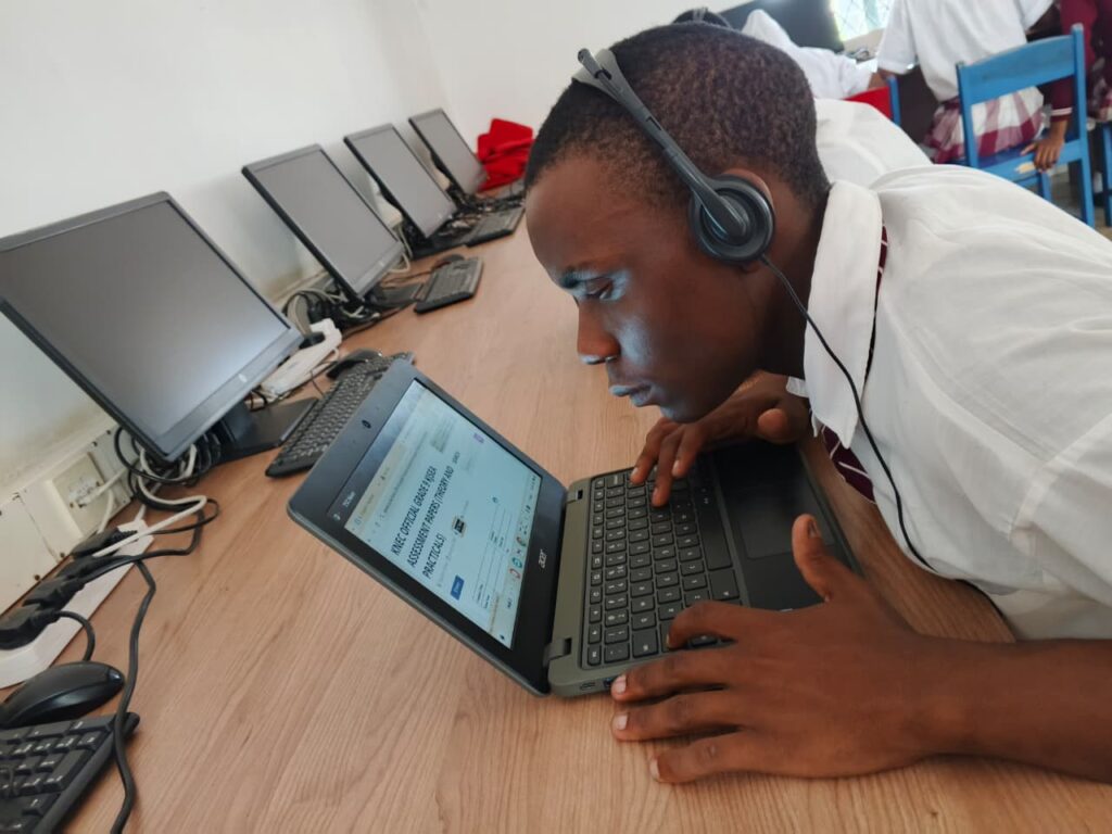 A student wearing headphones leans very close to a laptop screen while working in a computer lab. Several desktop computers are arranged on the desk beside him, and other students in school uniforms are seated in the background.