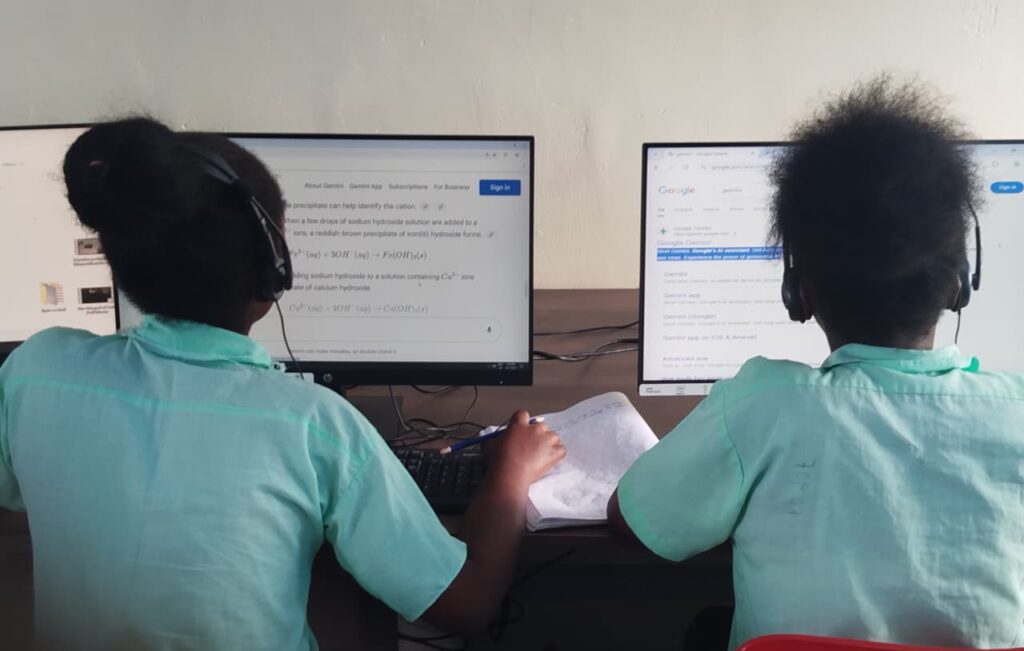 Two students wearing headphones sit side by side at desktop computers in a classroom. They are focused on the screens while one writes notes in a notebook placed on the desk.