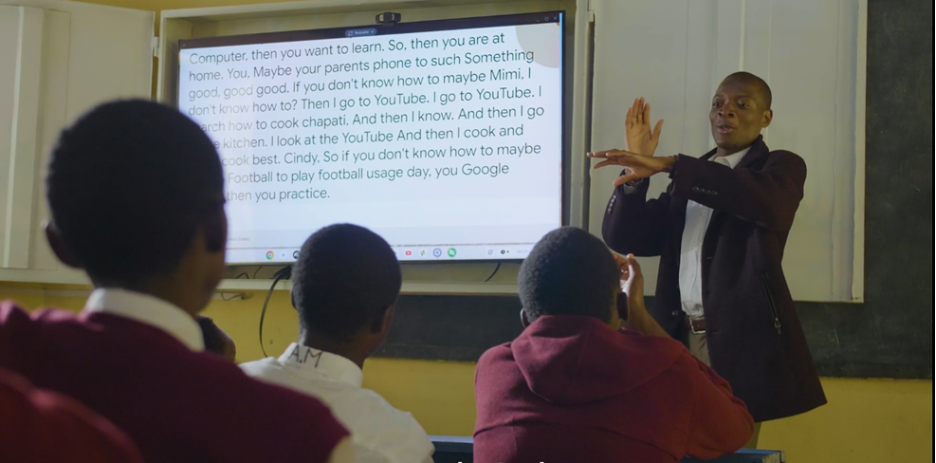 A teacher, a person with short hair wearing a dark blazer over a collared shirt, stands in a brightly lit classroom, gesturing with their hands towards a projected screen behind them. The screen displays large white text describing how to use a computer or phone to search and learn. Three students, seen from behind in maroon and white uniforms, are seated at desks, looking at the teacher and the screen. 