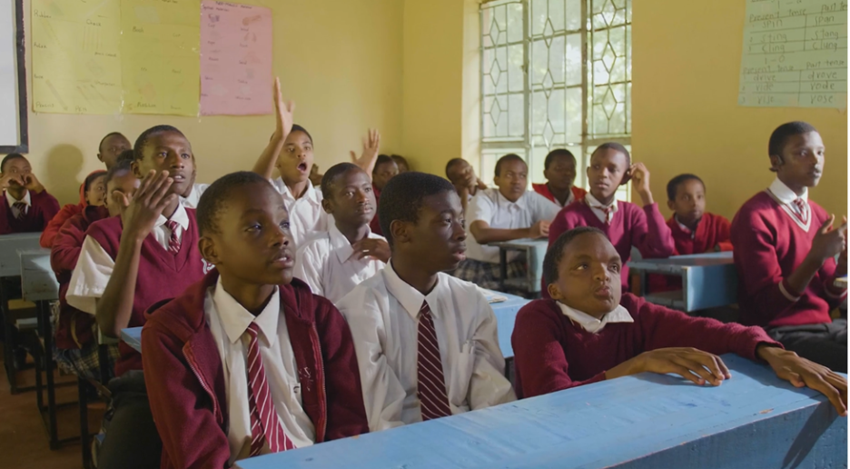 A classroom scene showing a group of students, mostly male, seated at light blue wooden desks. They are all wearing maroon and white school uniforms. Several students are actively engaged, with one raising their hand high and others looking intently towards the front of the classroom. Natural light streams in through a large window on the right.