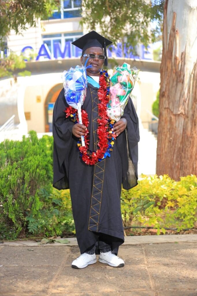 Maurice is wearing a black graduation gown and cap, adorned with colorful celebratory garlands around the neck. The person is holding two large wrapped bouquet gifts—one blue and one multicolored. They are wearing sunglasses and white sneakers. Behind them is a campus building entrance partially visible, with trees and greenery surrounding the area.