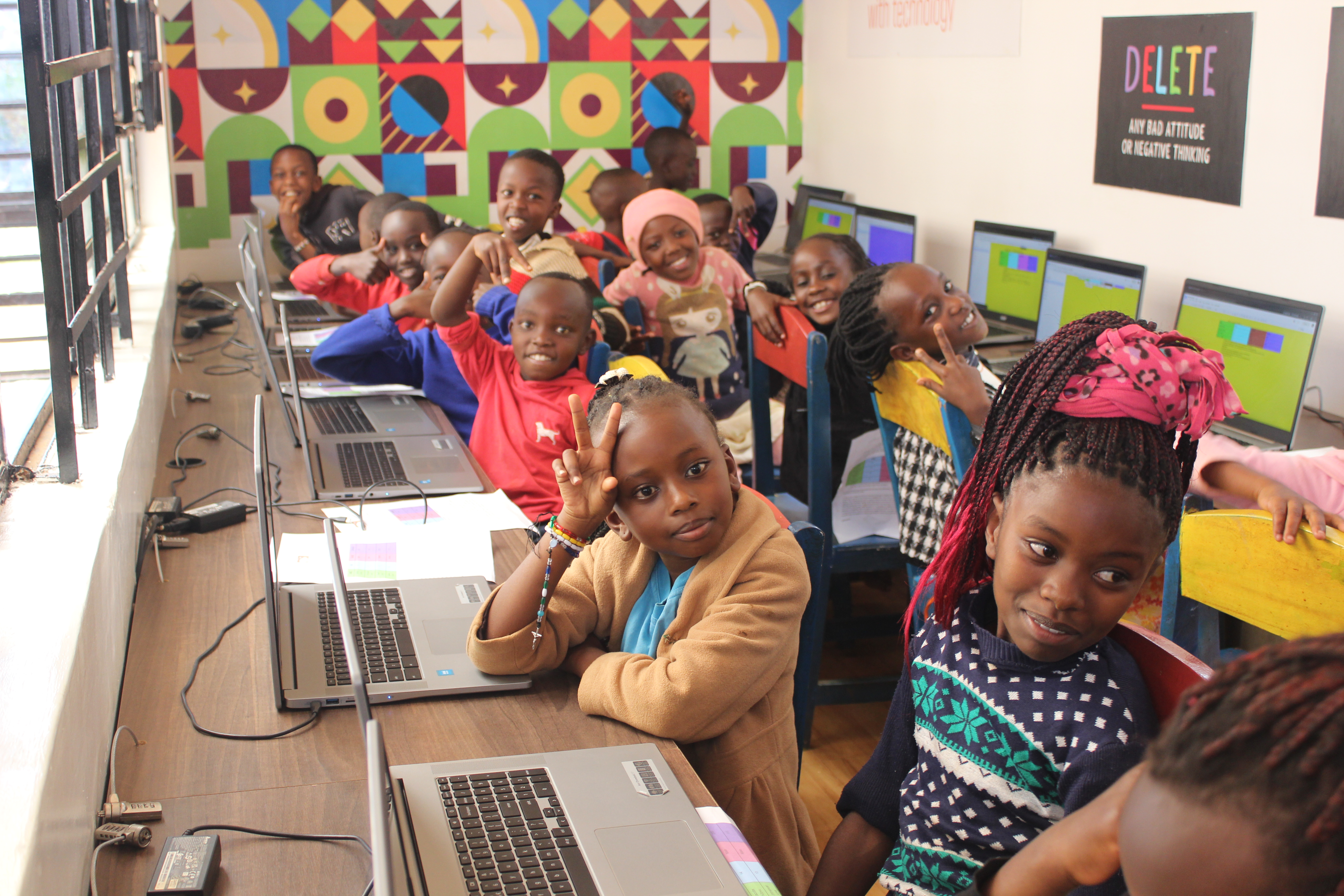 A group of smiling schoolchildren sit in a computer lab, each with a laptop open on long desks. The children face the camera, some flashing peace signs, while colorful geometric wall art and desktop screens are visible in the background, creating a lively and engaging learning environment.