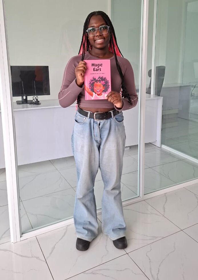 A picture of Jasmine with long braided hair and glasses stands indoors in a bright office with glass walls, smiling while holding up a pink children’s book titled Magic Ears. She is wearing a long-sleeve top, light blue jeans, and black shoes, with desks and computer monitors visible in the background.