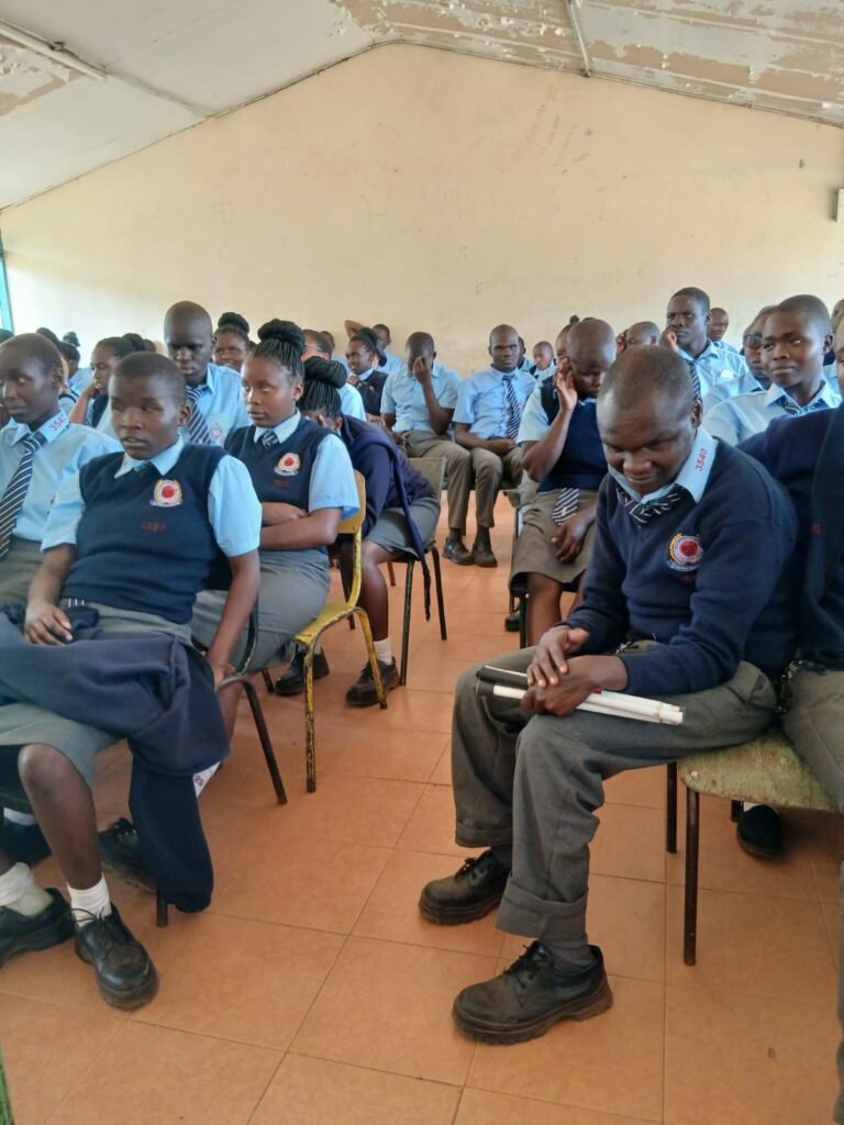 Secondary school students in blue uniforms sit closely together on chairs in a classroom during a session. In the foreground, a student holds a white mobility cane while seated, with other learners behind them listening quietly. The room has tiled floors, plain walls, and a worn ceiling, capturing a focused learning environment.