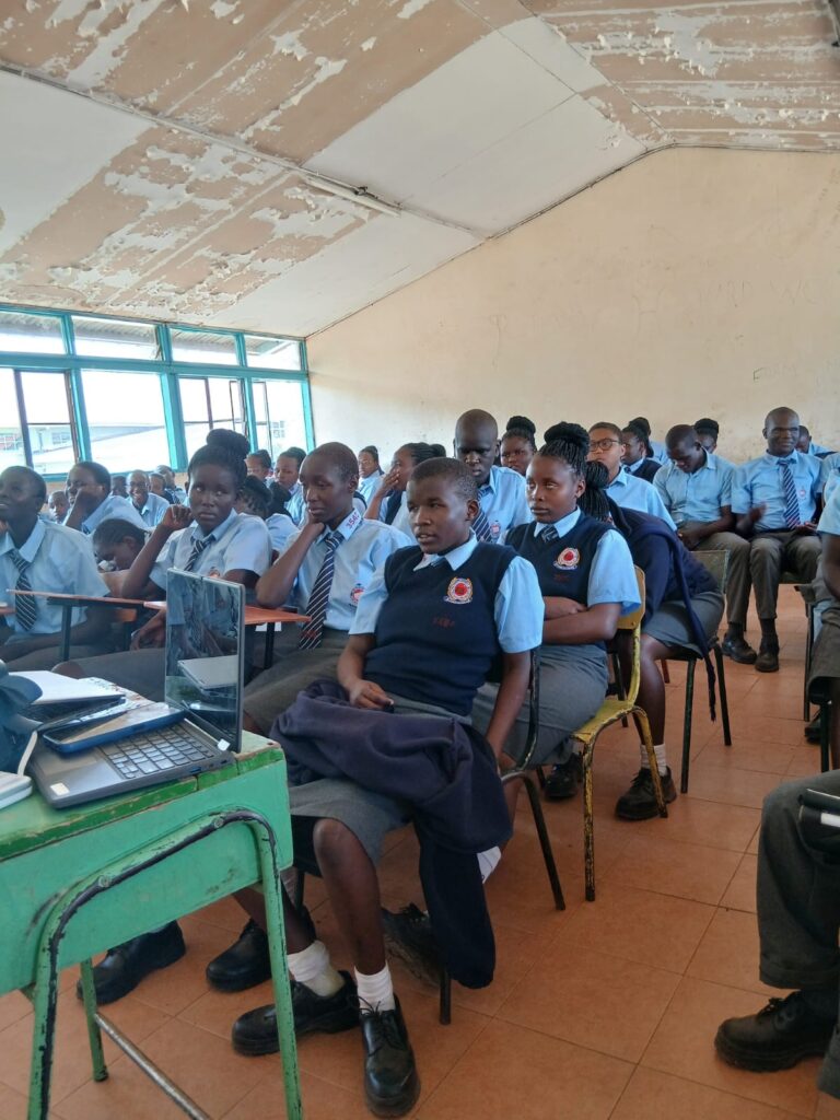 A classroom filled with secondary school students wearing blue uniforms, seated closely together on chairs and desks while attentively listening to a presentation. A laptop sits on a small green desk at the front, suggesting a lesson or talk in progress. The room has large windows on one side letting in natural light, and a slightly worn ceiling, indicating a well-used school learning space.