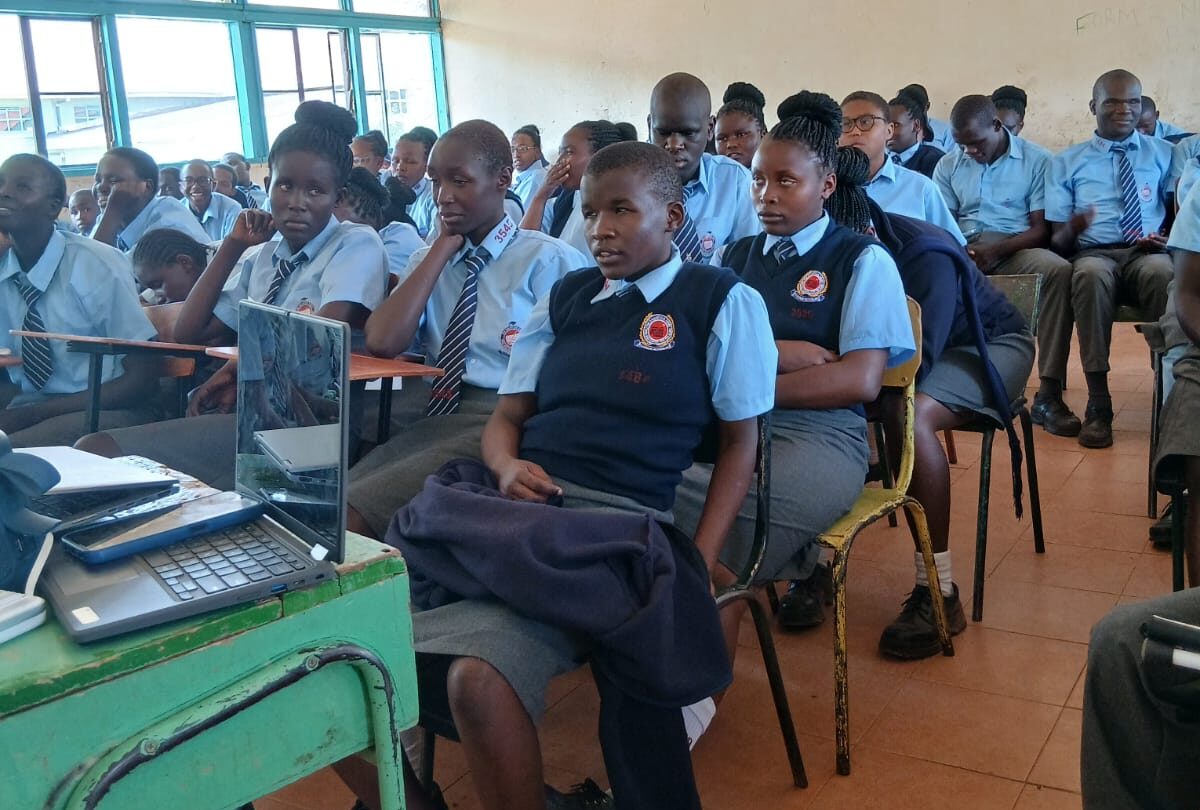 A classroom filled with secondary school students wearing blue uniforms, seated closely together on chairs and desks while attentively listening to a presentation. A laptop sits on a small green desk at the front, suggesting a lesson or talk in progress. The room has large windows on one side letting in natural light, and a slightly worn ceiling, indicating a well-used school learning space.