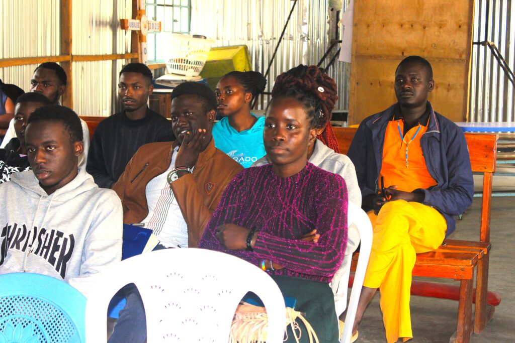 A group of participants sits indoors on plastic chairs and wooden benches during a training or workshop session. Attendees face forward and listen attentively to a speaker outside the frame. The meeting takes place in a hall with corrugated metal walls, wooden framing, and natural light coming through windows. Some participants hold notebooks or folders, while others sit with hands folded, focused on the presentation.