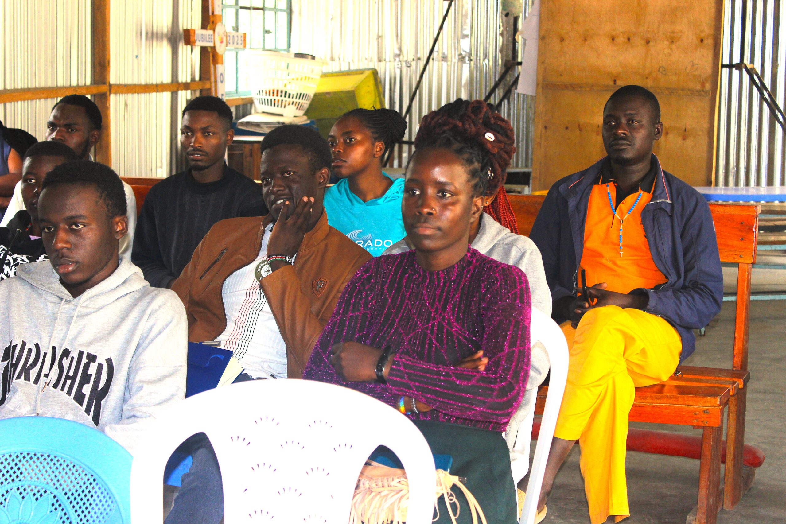 A group of participants sits indoors on plastic chairs and wooden benches during a training or workshop session. Attendees face forward and listen attentively to a speaker outside the frame. The meeting takes place in a hall with corrugated metal walls, wooden framing, and natural light coming through windows. Some participants hold notebooks or folders, while others sit with hands folded, focused on the presentation.