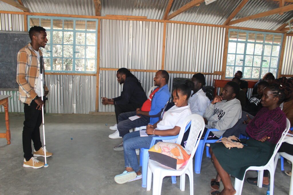 Julius stands at the front of a classroom-style training session, addressing a group of participants seated on plastic chairs and taking notes. He is using a white mobility cane. The room has corrugated metal walls, large windows, a chalkboard, and a concrete floor. Participants face the speaker attentively in an informal learning environment.