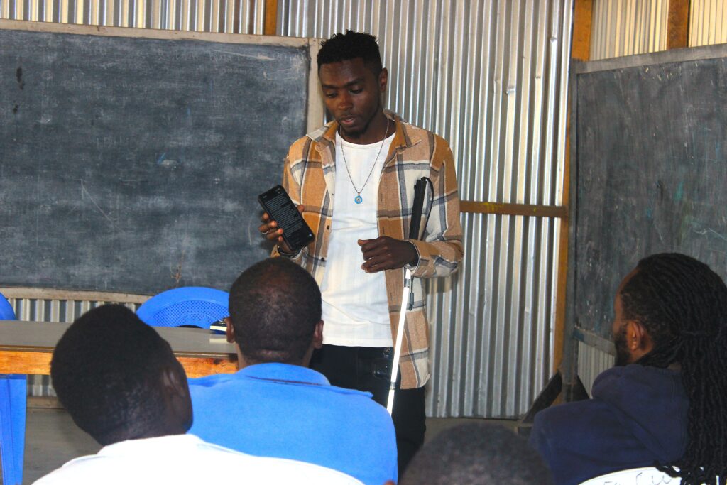 Julius stands at the front of a training room, demonstrating accessibility features on a smartphone to a group of seated participants. He holds a white mobility cane and speaks from between two chalkboards mounted on corrugated metal walls. Several participants are visible in the foreground, facing the presenter and listening during the workshop. The room contains simple tables and plastic chairs, creating a practical community learning environment.