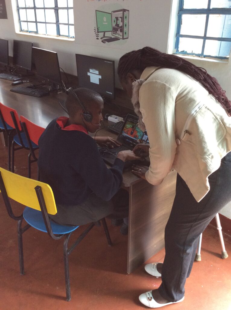 Janet is wearing a headset sits at a laptop in an inABLE computer lab while receiving one-on-one guidance from Valarie, an instructor. The instructor leans over the workstation, pointing at the laptop screen as they work together on a digital task. Desktop computers line the long table beside them, and large windows provide natural light to the room. Educational technology graphics are displayed on the wall, highlighting a supportive and inclusive environment for digital skills training and technology-assisted learning.