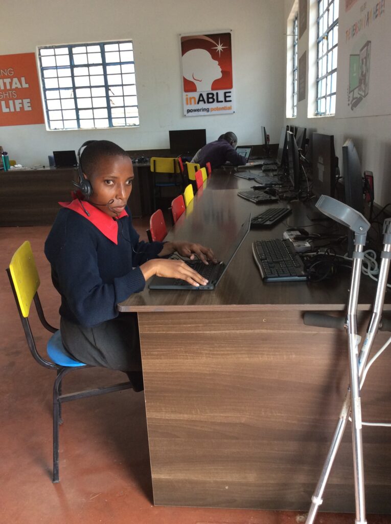 Janet sits at a computer workstation in an inABLE computer lab, using a laptop with a headset and microphone. Several desktop computers are arranged along a long table, while another learner works at a computer in the background. The room is well lit by large windows, and an inABLE logo sign is displayed on the wall. A pair of forearm crutches is positioned beside the workstation, highlighting the accessible learning environment and the use of technology for digital skills development.